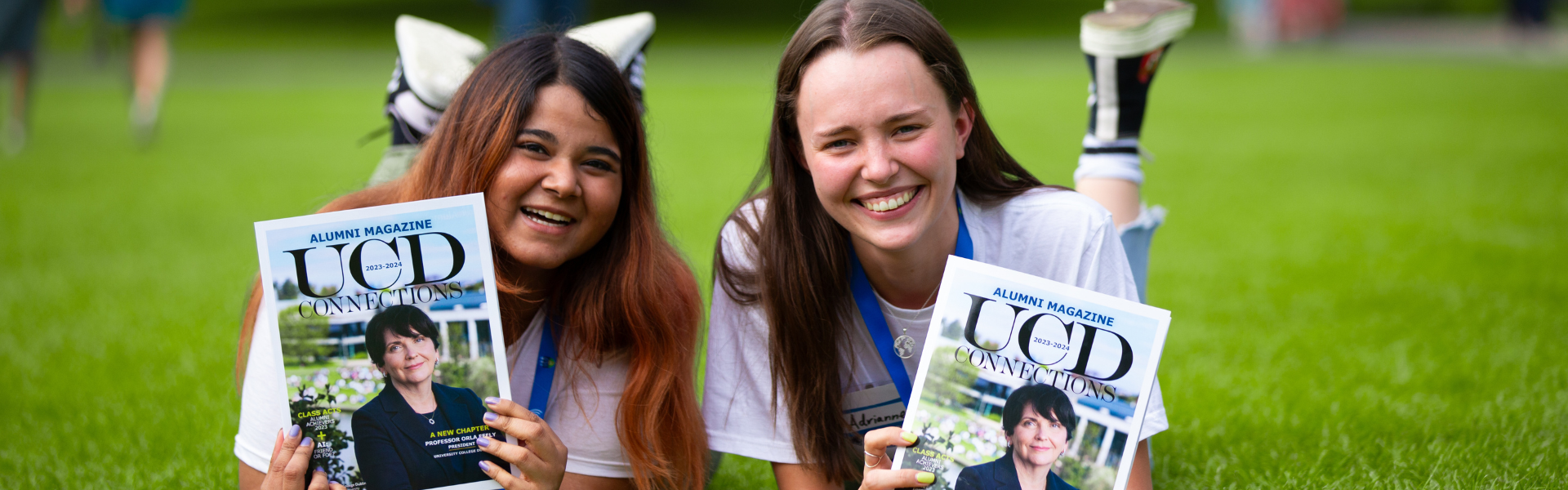 Two UCD alumnae lying on grass holding up UCD Connections, the annual alumni magazine.
